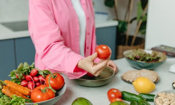 Person in pink overshirt holding a fresh tomato with various organic vegetables on a countertop.